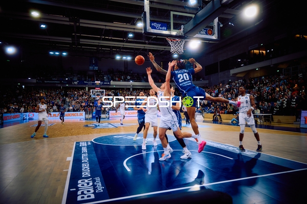 Ein Spieler von Brose Bamberg mit der Nummer 20 springt zum Korb für einen Wurf, während zwei Verteidiger von MLP Academics Heidelberg versuchen, ihn zu blocken.