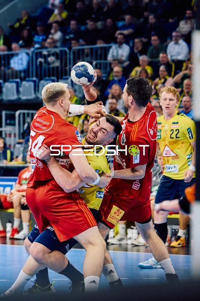 Ein Spieler der Rhein-Neckar Löwen im gelben Trikot ist im Zweikampf mit zwei Spielern der TSV Hannover Burgdorf, die das rote Trikot tragen. Einer der Rhein-Neckar Löwen-Spieler mit der Nummer 22 scheint sich durch die gegnerische Abwehr zu kämpfen.