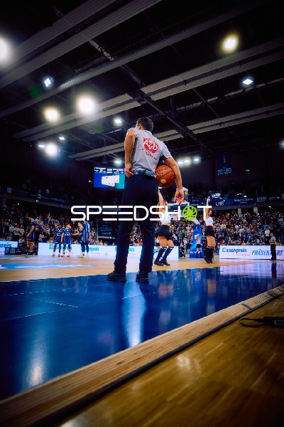 Ein Schiedsrichter steht an der Seitenlinie mit einem Basketball in der Hand, während das Spiel im Hintergrund läuft.