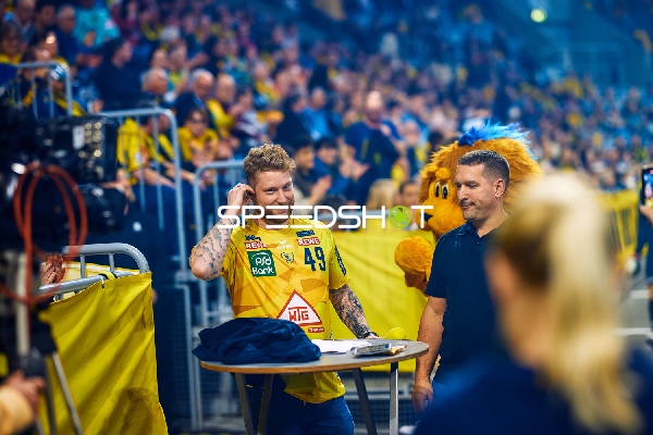 Kevin Gerwin steht in der Nähe eines Tisches am Spielfeldrand. Ein Maskottchen ist ebenfalls zu sehen.