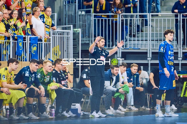 Die Trainerbank während eines Handballspiels mit Spielern und Trainern, die aktiv das Spielgeschehen verfolgen.