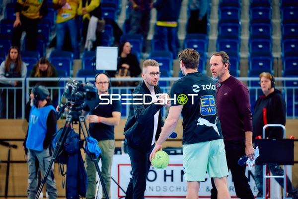 Interaktion zwischen einem Handballspieler und TV-Personal am Spielfeldrand.