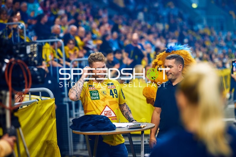 Kevin Gerwin steht in der Nähe eines Tisches am Spielfeldrand. Ein Maskottchen ist ebenfalls zu sehen.