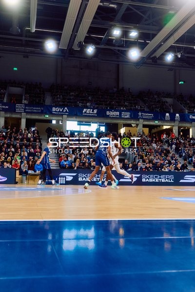 Basketballspiel in Aktion mit einem Spieler im blauen Trikot, der gegen einen Spieler im weißen Trikot verteidigt.