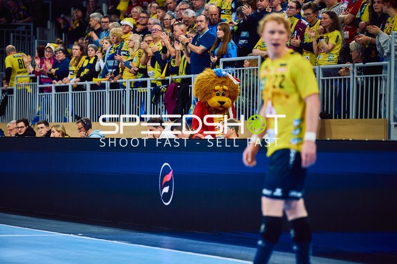 Ein Spieler von Rhein Neckar Löwen auf dem Spielfeld, mit Fans im Hintergrund, darunter ein Maskottchen.
