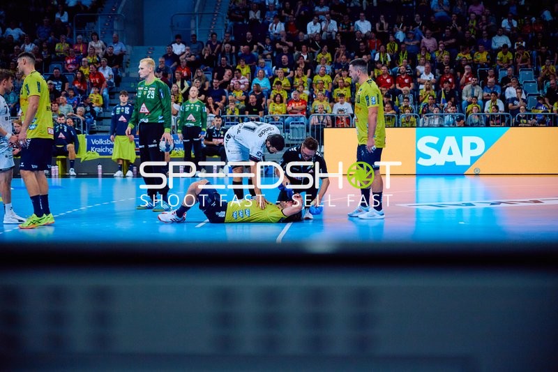 Ein Spieler von Rhein Neckar Löwen liegt verletzt auf dem Spielfeld, während andere Spieler und Betreuer sich um ihn kümmern.