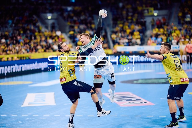 Ein Spieler von Frisch Auf Göppingen springt für einen Wurf hoch, während zwei Spieler von Rhein-Neckar Löwen versuchen ihn zu blocken.