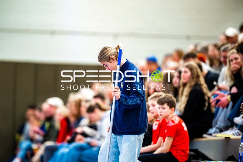 Eine Person reinigt den Boden in einer Sporthalle, während Zuschauer auf der Tribüne sitzen.