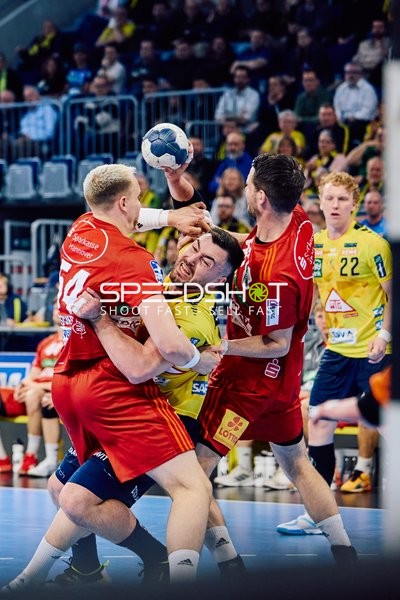 Ein Spieler der Rhein-Neckar Löwen im gelben Trikot ist im Zweikampf mit zwei Spielern der TSV Hannover Burgdorf, die das rote Trikot tragen. Einer der Rhein-Neckar Löwen-Spieler mit der Nummer 22 scheint sich durch die gegnerische Abwehr zu kämpfen.