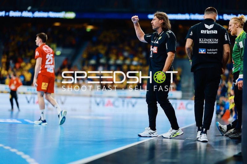 Ein Trainer der Rhein-Neckar Löwen gibt Anweisungen am Spielfeldrand, während Tilen Strmljan (Nr. 25) von TSV Hannover Burgdorf auf dem Spielfeld zu sehen ist.